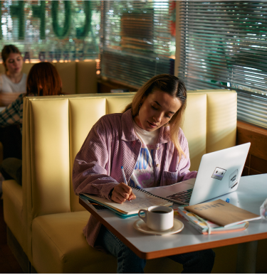 A young woman sitting at a diner with a laptop and writing in her notebook